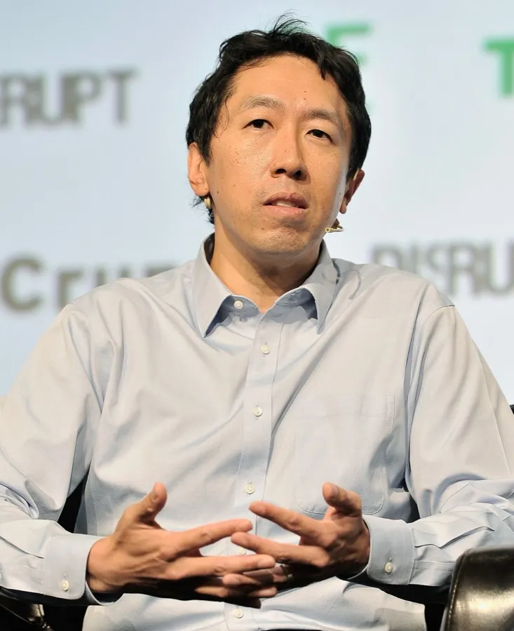 Andrew Ng speaking on stage, hands together, in a light blue shirt.
