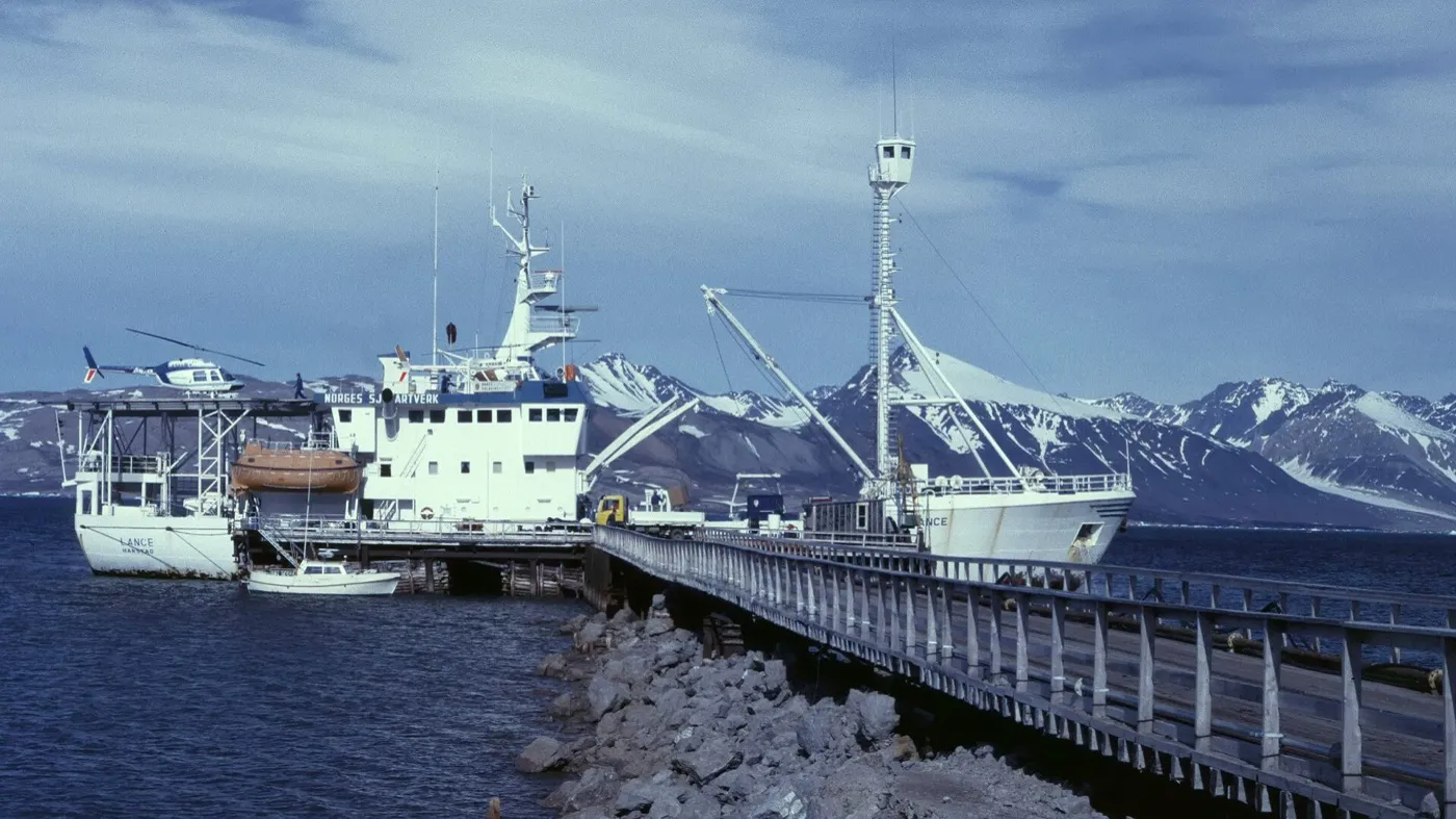 A research ship with a helicopter on deck, moored at a long pier in dark blue water, with snow-capped mountains behind.