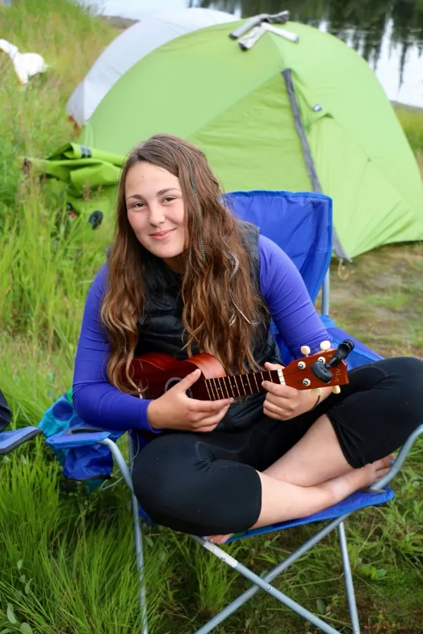 A young woman smiling in a camp chair outdoors, holding a ukulele, with a green tent behind her.