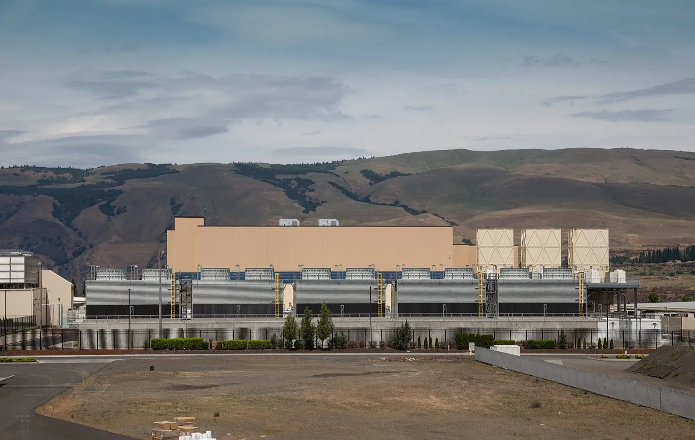 A large beige data centre building with rows of grey cooling units in front and dry brown hills behind, under a cloudy sky.